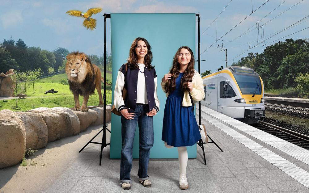 Elise und Sophie am Bahnsteig in Wuppertal Eine junge Frau und ein Mädchen stehen am Bahnsteig Wuppertal vor einer türkisen Leinwand. Links ist ihr Reiseziel, der Zoo in Wuppertal zu sehen und rechts ein gelb-weißer Zug der Linie RE 13 von der eurobahn..